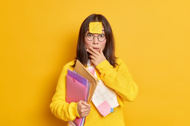 Photo of puzzled surprised Asian female student has memo note stuck on forehead prepares coursework carries folders with papers makes education project works at her assignment studies remotely