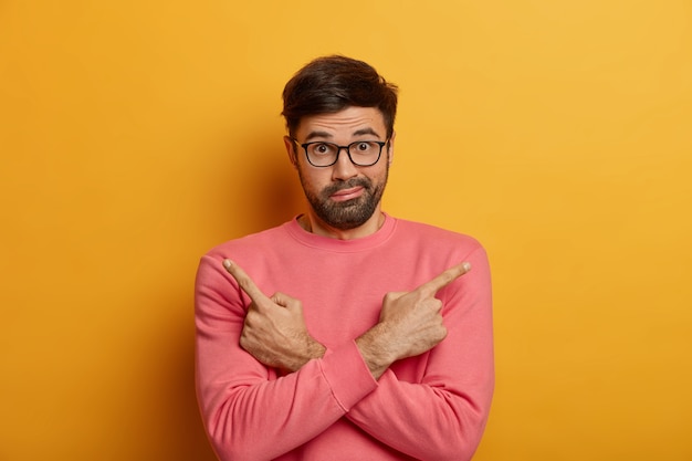 Photo of puzzled unshaven man points sideways, crosses arms over chest, doubts between two choices or variants, has bewildered look, wears spectacles and rosy sweater, isolated on yellow wall