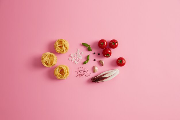 Photo of raw pasta nests with ingredient to cook. Red salad chicory, cherry tomatoes, basil, garlic and dried chili pepper threads on rosy background. Preparing delicious macaroni. Italian cuisine