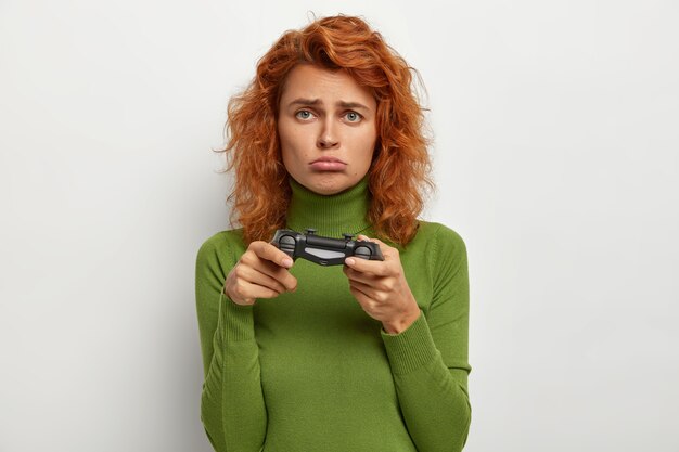 На фото рыжеволосая девочка-подросток играет с джойстиком, с недовольным выражением лица, проигрывает в видеоигре, проводит свободное время дома, будучи настоящим геймером. Люди, досуг, концепция развлечений
