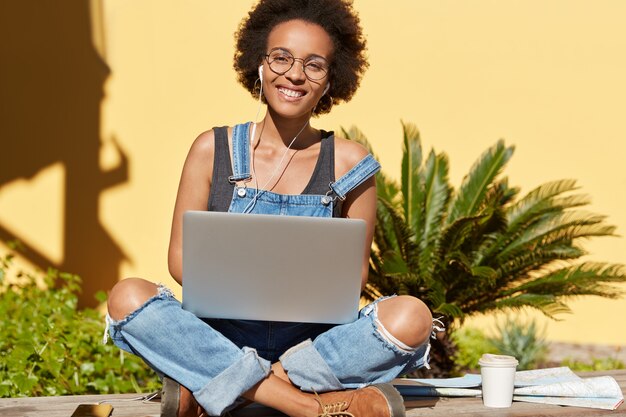 Photo of relaxed carefree dark skinned woman sits crossed legs, surfes internet web site on laptop computer, enjoys fresh drink, listens pleasant audio track with earphones. African American blogger