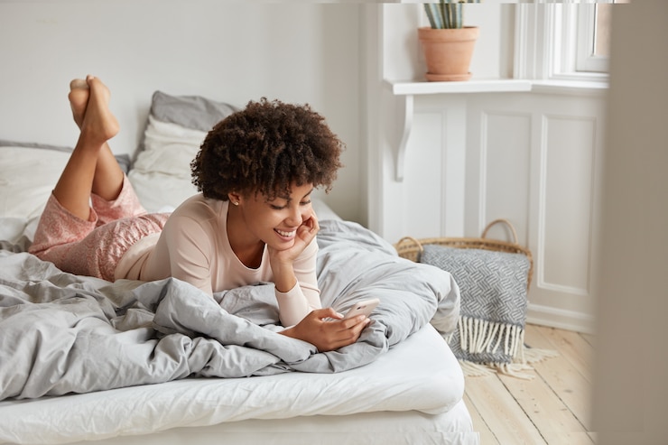 Free Photo photo of relaxed dark skinned woman watches online story on mobile phone, reads post with exciting information, dressed in casual home clothes, lies on bed, has happy expression. cozy interior