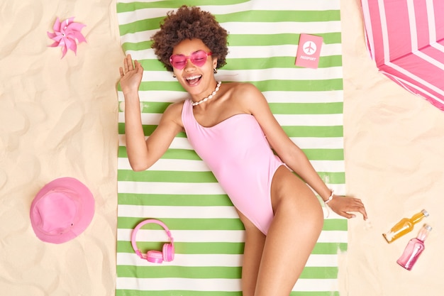 Photo of relaxed glad young afro american model smiles pleasantly wears pink sunglasses and bikini lies on green striped towel surrounded by necessary items sunbathes at beach on white sand