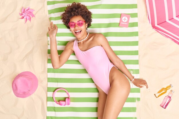 Photo of relaxed glad young Afro American model smiles pleasantly wears pink sunglasses and bikini lies on green striped towel surrounded by necessary items sunbathes at beach on white sand