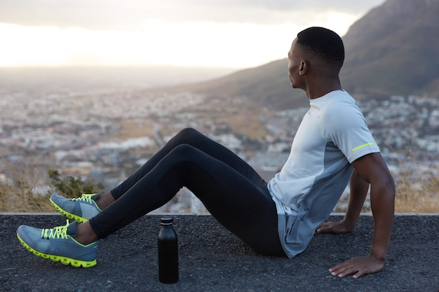 Photo of relaxed man with short hair, dark skin, sits on highway with bottle of cold water, wears tracksuit, focused aside, enjoys spare time, mountain view, rests after workout. Fitness, exercising