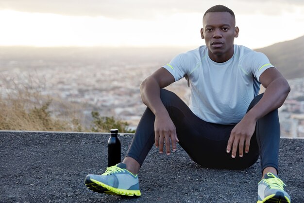 Photo of relaxed masculine guy with healthy dark skin, wears sportsclothes, drinks water, sits at road above daylight and clear sky takes break after running exercises. Relaxation, sport