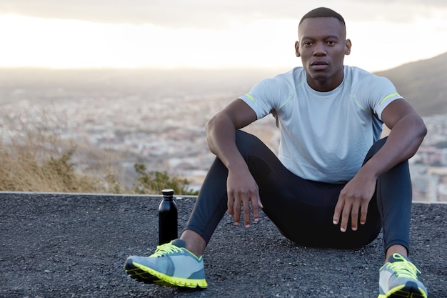 Photo of relaxed masculine guy with healthy dark skin, wears sportsclothes, drinks water, sits at road above daylight and clear sky takes break after running exercises. Relaxation, sport