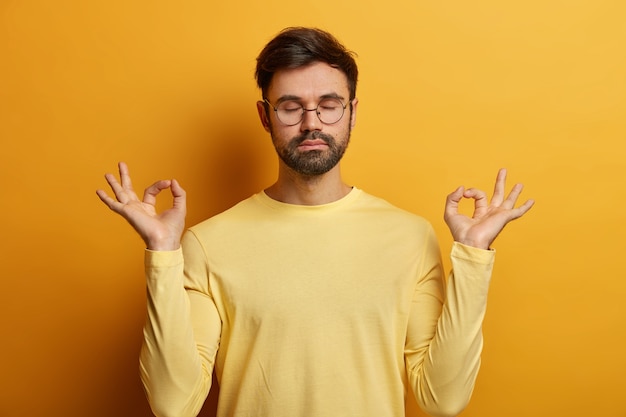 Photo of relaxed unshaven European man stands in lotus pose, makes zen gesture, breathes deeply and tries to relax, keeps eyes closed, wears glasses and jumper, poses indoor, reaches nirvana