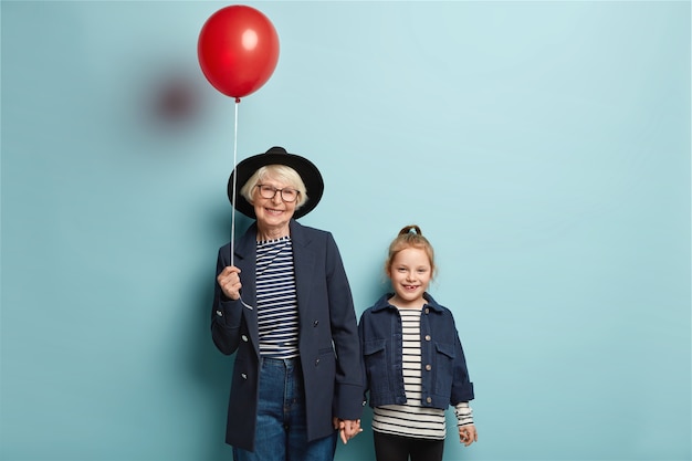 Photo of retired senior woman holds hand of little adorable granddaughter, come on party, hold red balloon