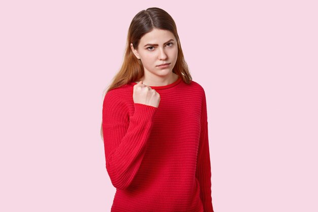 Photo of sad angry young European woman with straight dark hair, shows fist at camera, dressed in red jumper, poses over rosy wall expresses threating, reproaches and promises to revenge.