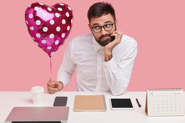 Photo of sad bearded Caucasian man in formal clothes, carries valentine, feels lonely, has no love, dreams about new relationship, sits at desktop