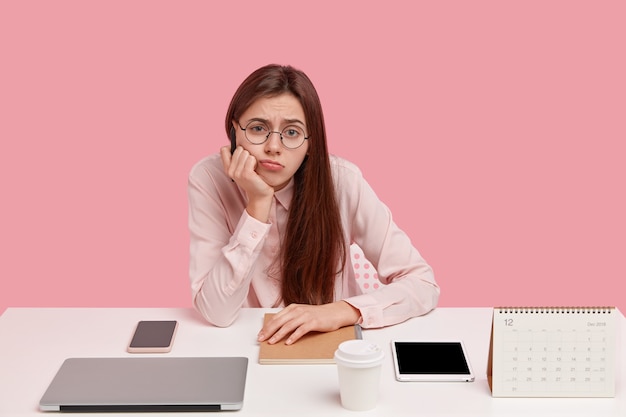 Free photo photo of sad european woman holds chin, looks in discontent, wears round spectacles and elegant shirt, doesnt want to work