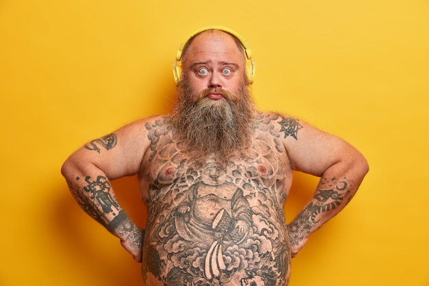 Photo of sad plump bearded man looks with surprised expression listens favorite song in headphones, keeps hands on hips, has big fat tummy, tattooed body, isolated on yellow wall