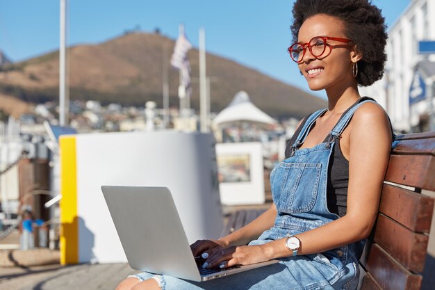Photo of satisfied copywriter or freelancer keyborads information on laptop keyboard, thinks about something creative, wears casual clothes, models at bench outdoor, works freelance, uses 4G