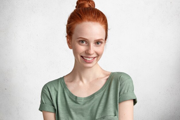 Photo of satisfied freckled adorable young woman with ginger hair tied in knot, smiles pleasantly at camera, rejoices coming holidays, isolated over white