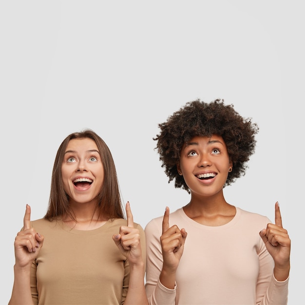 Photo of satisfied mixed race women with joyful expression indicates upwards with both index fingers, focused up
