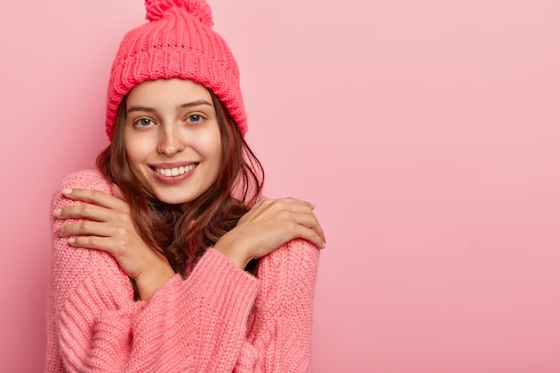 Photo of a satisfied smiling woman being warm in knitted winter sweater, crosses arms over chest and touches shoulders, has appealing look, poses against pink background, free space aside