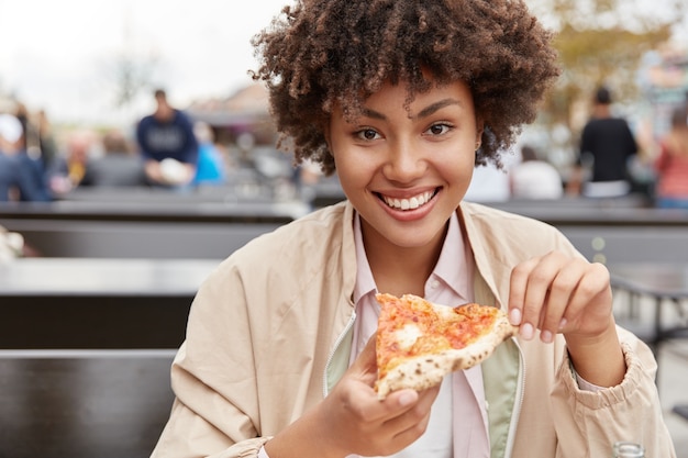 Photo of satisfied teenage girl with dark healthy skin, enjoys delicious meal, holds piece of pizza