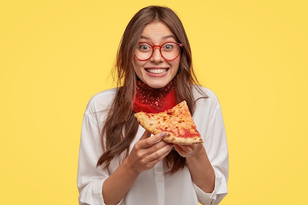 Photo of satisfied woman holds piece of pizza, feels pleased as spends free time with friends in pizzeria, looks happily directly  wears casual outfit, isolated over yellow wall. Lunch