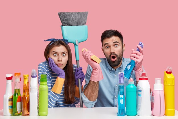 Photo of scared astonished lady and guy have surprised expressions, look with stupor, carry detergents, sponge, brush