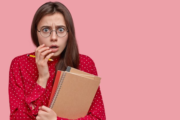 Photo of scared Caucasian lady looks in despair, opens mouth from surprise, carries spiral notepad and pencil