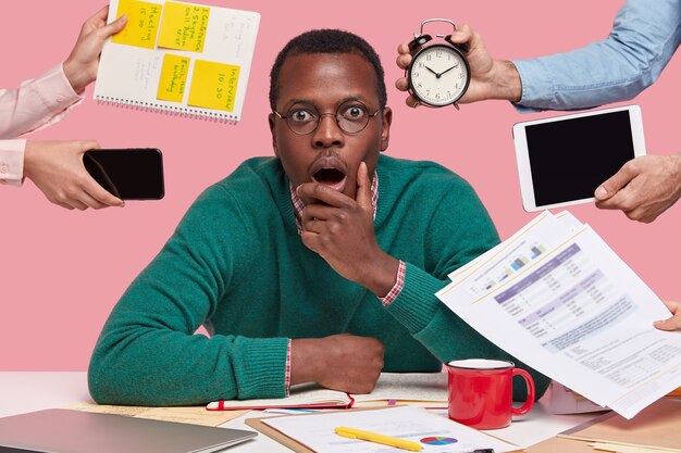 photo of scared dark skinned guy holds chin, opens mouth widely, wears glasses, surrounded with peoples hands holding electronic gadgets