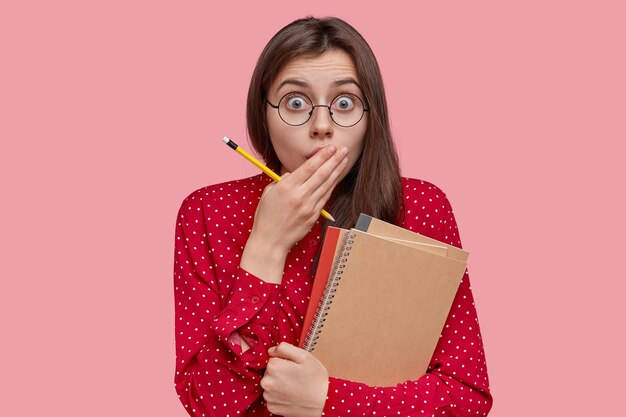 Photo of scared young woman with dark hair covers mouth, has surprised expression, uses notepad for writing records