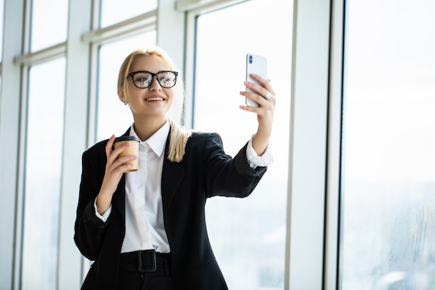 Фотография секретарши в торжественной одежде, держащей кофе на вынос в руке и делающей селфи на мобильном телефоне в офисе