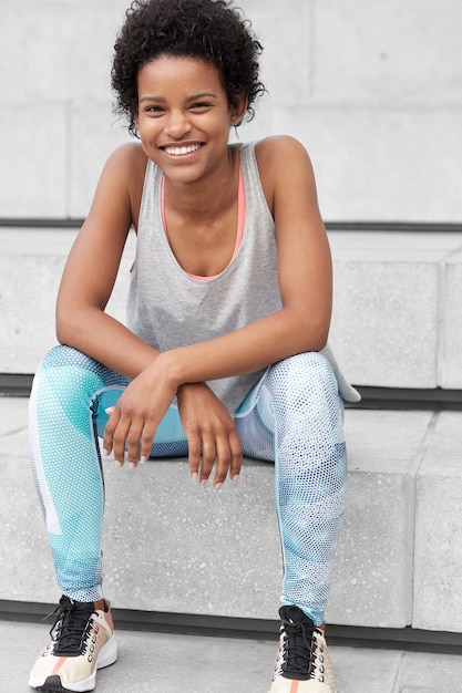 Photo of self confident black lady in sportsclothes, has happy relaxed expression, smiles broadly, being glad to win sport competitions, sits at stairs, ready to achieve new goals. Ethnicity, sport