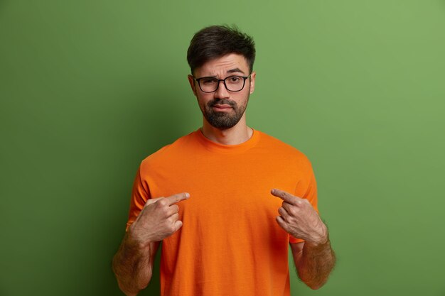 Photo of self confident cheeky sassy hipster points at himeself, says you may rely on me, wears glasses and orange t shirt, isolated on green wall. Assertive arrogant bearded man indoor