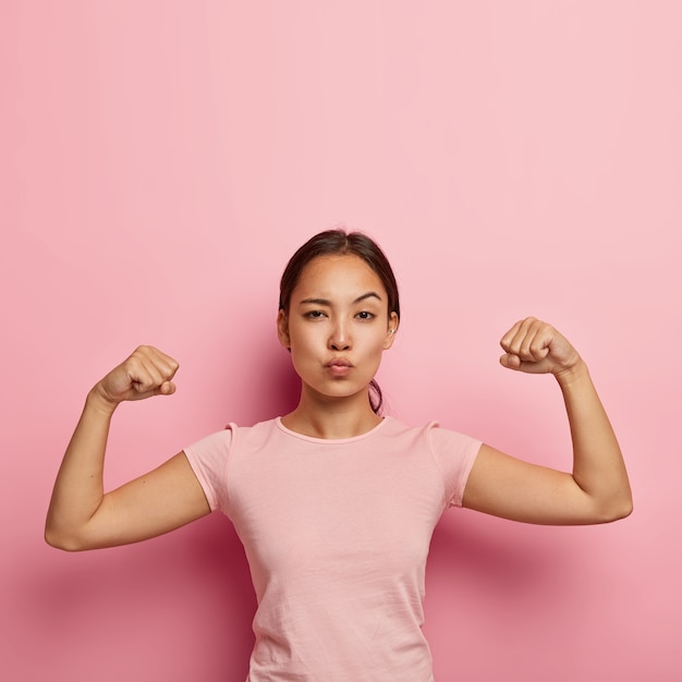 La foto della ragazza asiatica seria sicura di sé tiene le labbra piegate, mostra i suoi muscoli e la sua forza, non indossa trucco, indossa una maglietta casual, isolata sul muro rosa con copia spazio sopra per informazioni