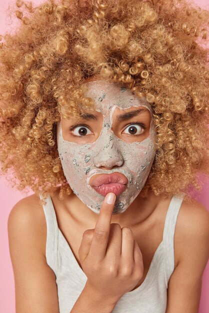 Фотография серьезной внимательной молодой женщины с вьющимися густыми волосами, надутыми губами, наносит питательную косметическую маску для ухода за кожей, одетая в повседневную футболку, позирует в помещении Уход за лицом и концепция спа