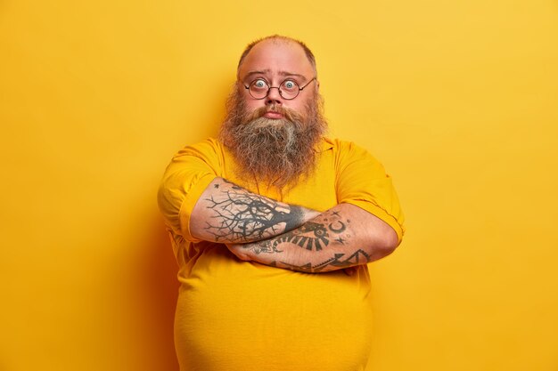 На фото серьезный бородатый мужчина стоит со скрещенными руками, с большим пивным животом, озадачен неудачной диетой, имеет лишний вес из-за неправильного питания, выглядит удивленно, стоит в помещении