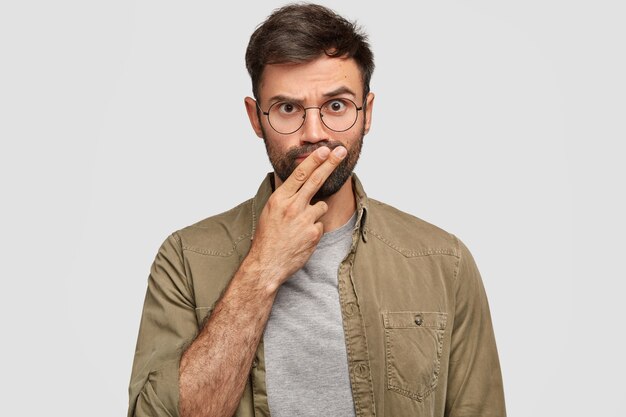 На фото серьезный бородатый молодой парень держит два пальца во рту, пытается принять решение, смотрит со строгим выражением лица, собирается кого-то наказать, носит повседневную одежду, изолирован на белой стене
