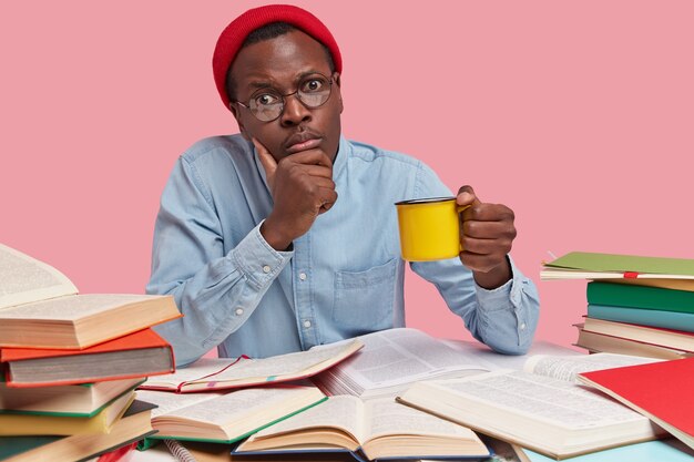 Photo of serious black man holds chin, carries yellow mug of drink, looks directly at camera, wears red hat and shirt