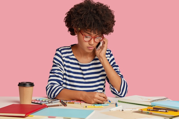 Photo of serious busy female worker with Afro hairstyle, creats illustration for project work, talks with partner via cellular, wears transparent glasses and striped clothes, isolated over pink wall