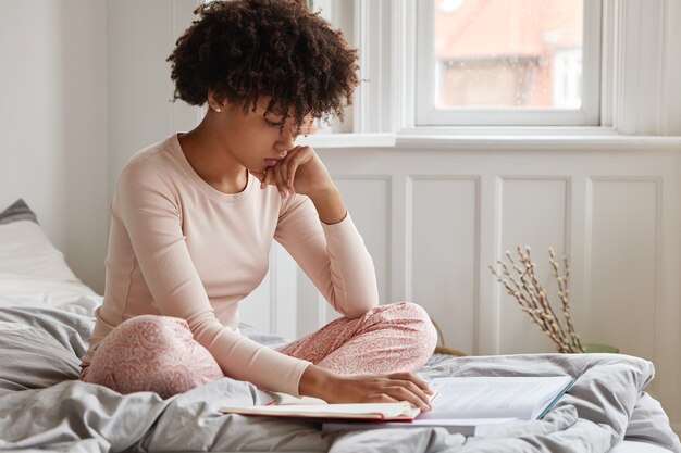 Photo of serious concentrated dark skinned young woman wears underwear, sits in lotus pose on bed, learns material from book and notebook,