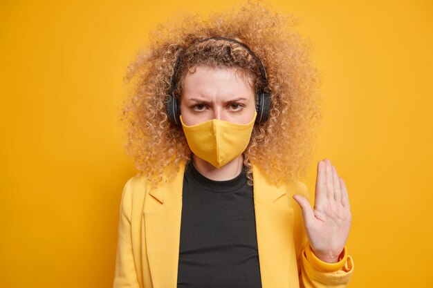 Photo of serious curly haired woman makes stop hand gesture to protect from Covid 19 asks to stop coronavirus outbreak and keeps social distancing listens music via headphones looks very strict