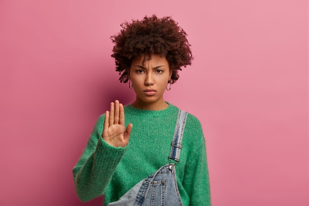 Photo of serious curly young woman rejects strange offer, pulls palm, declines proposal and looks in dissatisfaction , wears green sweater, warns not to pass any further