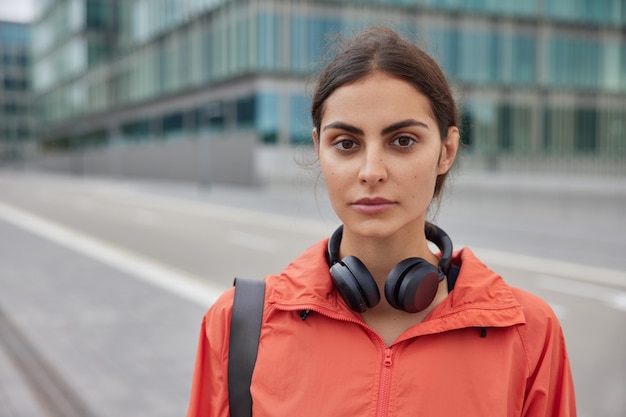 Photo of serious female sports trainer strolls on road alone wears jacket headphones around neck leads