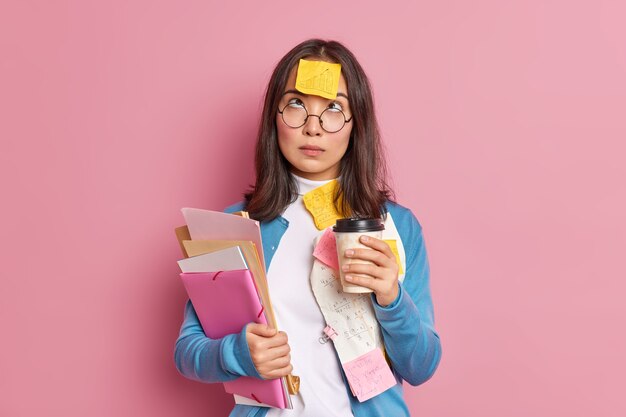 Photo of serious female student has coffee break concentrated above has reminding sticker stuck on forehead