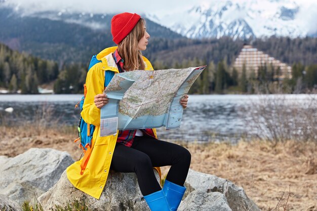 バックパックを持った真面目な女性旅行者の写真は、新しい目的地を探索し、石の上に座って地図を読み、場所を探します