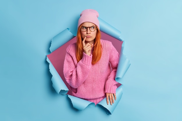 Photo of serious gloomy woman with red hair looks pensively aside wears pink hat knitted sweater.