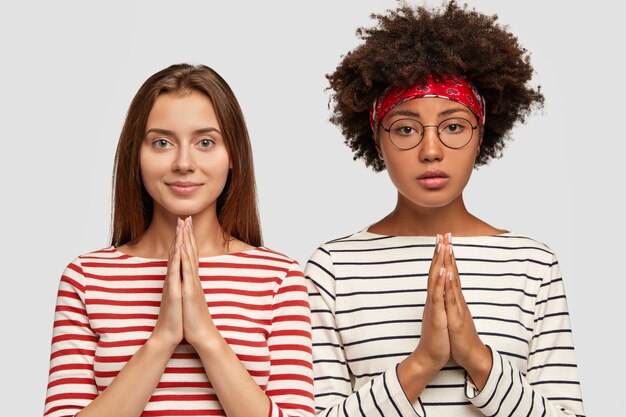 Photo of serious interracial woman have faithful expressions, keep palms pressed together