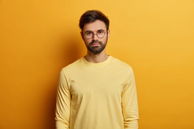 Photo of serious looking man has dark bristle, wears round spectacles and yellow jumper, direct gaze , poses indoor, has casual talk with someone. Monochrome. Face expressions concept