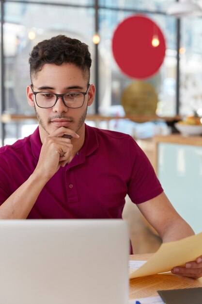 Photo of serious mixed race young man focused into monitor of modern laptop computer, installs application, updates software, makes distant job, holds chin, wears optical glasses for vision.