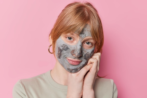 Photo of serious redhead girl leans on hands applies beauty clay mask on face for skin treatment has tender look at camera poses against pink background Facial care peeling and wellness concept