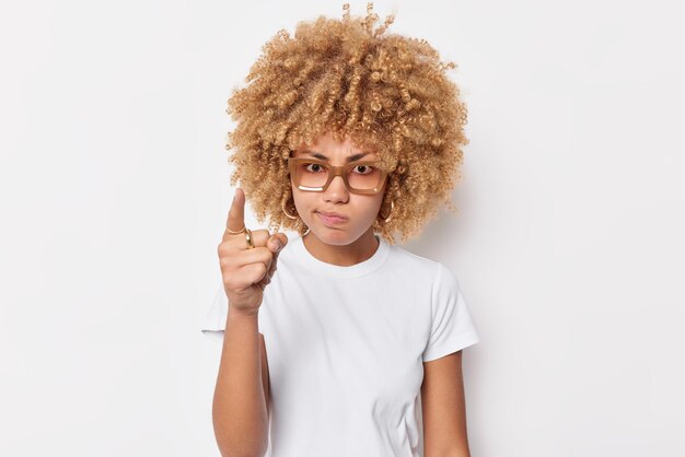 Photo of serious strict woman points index finger at camera warns you about something wears spectacles and casual t shirt isolated over white background. Angry girl says you must be attentive