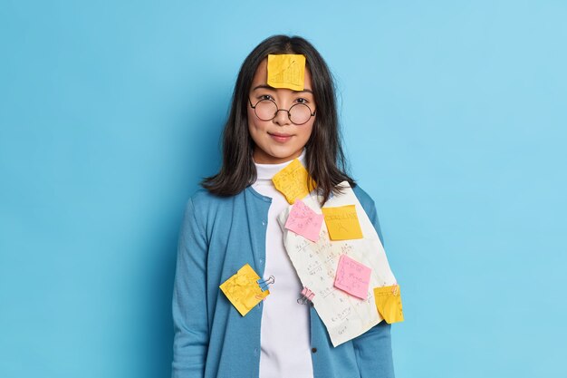 Photo of serious student makes notes on stickers and papers to remember information wears round spectacles prepares for private lesson works on coursework in university.