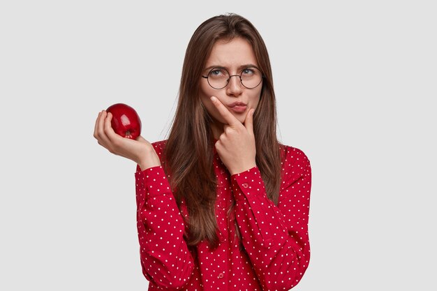 Photo of serious thoughtful pretty woman holds chin, carries apple, has contemplative expression, wears red shirt